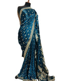 A teal blue silk saree with intricate gold and silver floral patterns and a wide gold border, draped on a black mannequin torso against a white fabric backdrop.