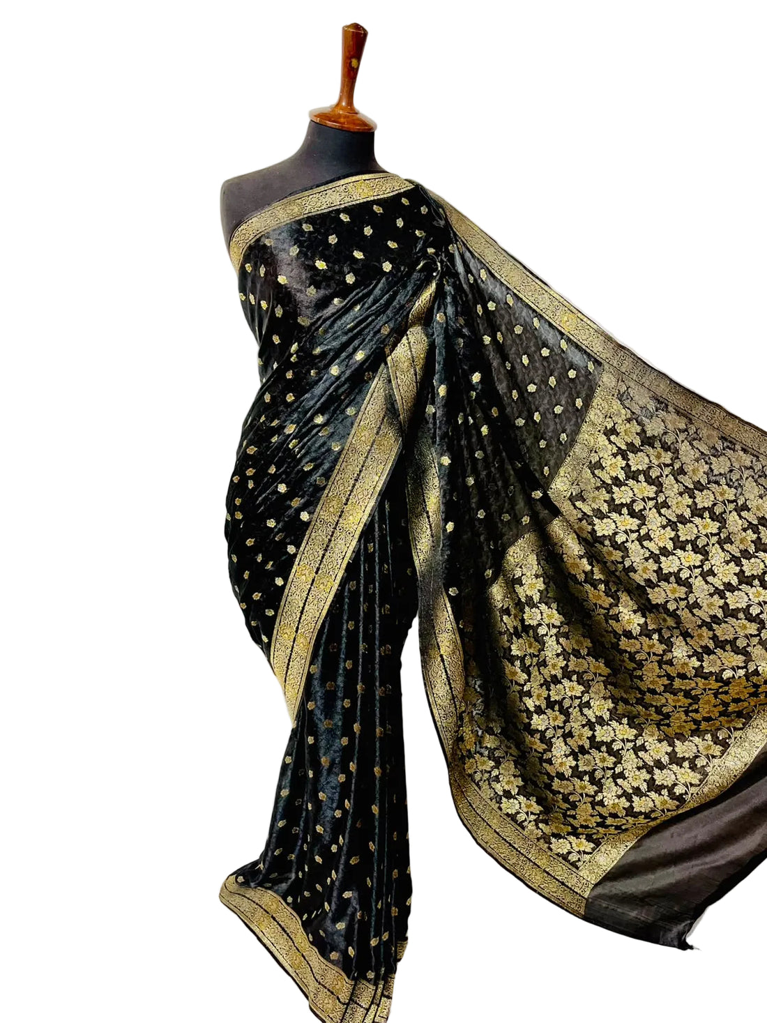A black silk saree with gold zari floral patterns and a gold border, draped on a black mannequin torso against a white fabric backdrop.