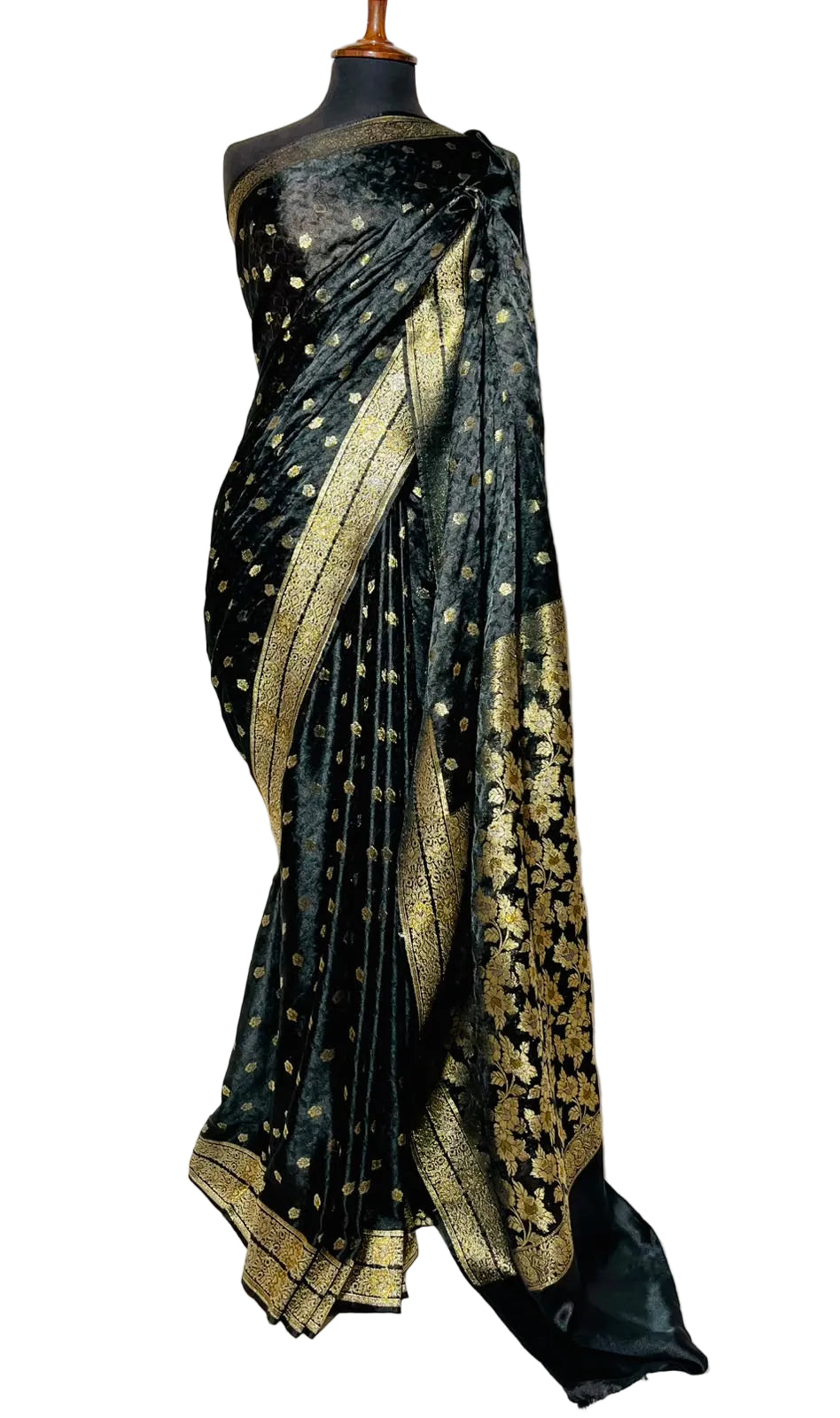 A black silk saree with gold zari floral patterns and a gold border, draped on a black mannequin torso against a white fabric backdrop.