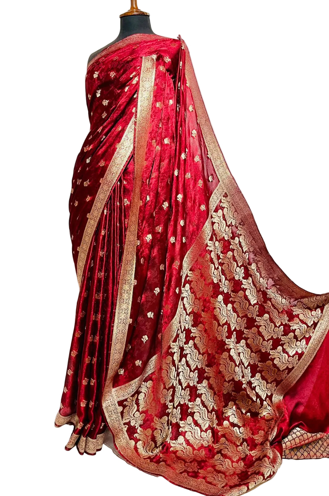 A deep red silk saree with intricate gold zari embroidery and a wide gold border, displayed on a black mannequin torso against a white fabric backdrop.