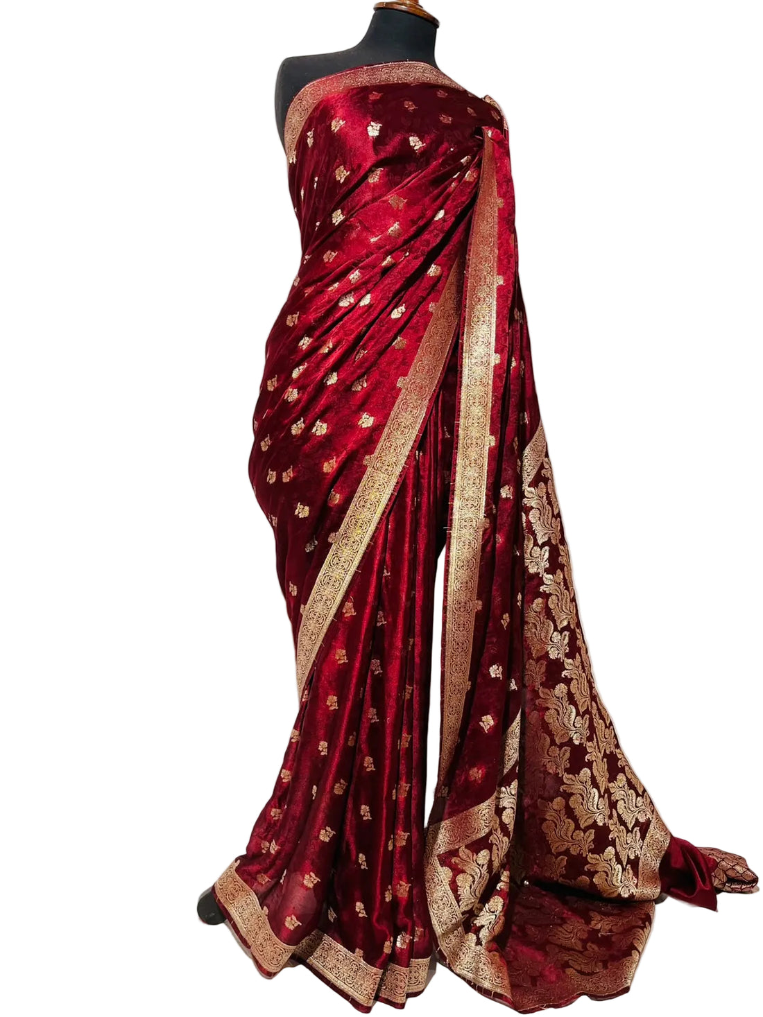 A deep red silk saree with intricate gold zari embroidery and a wide gold border, displayed on a black mannequin torso against a white fabric backdrop.