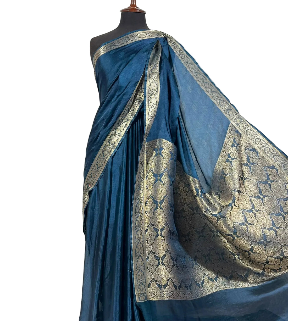 A blue silk saree with intricate gold zari brocade patterns draped on a black mannequin torso against a white fabric backdrop.