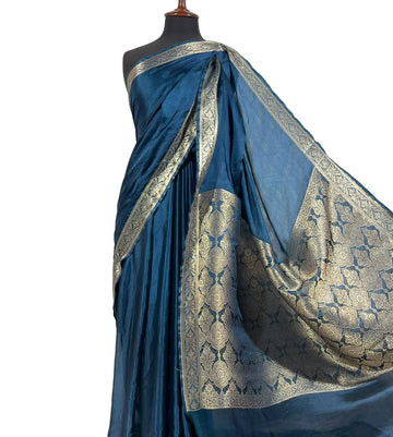 A blue silk saree with intricate gold zari brocade patterns draped on a black mannequin torso against a white fabric backdrop.